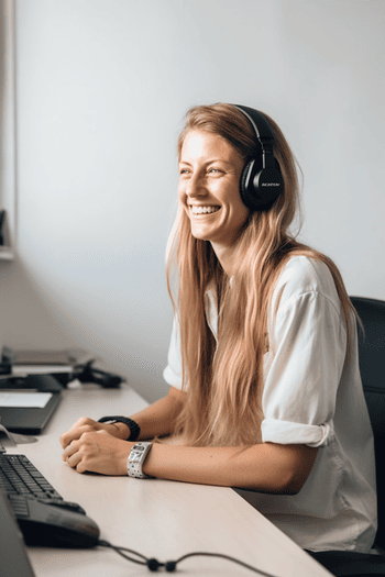 A white woman sitting at a desk, wearing a headset, and smiling while assisting a customer over the phone with a travel-related inquiry.